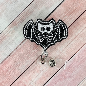 Può includere: Pipistrello ricamato in bianco e nero con glitter, attaccato a un porta badge in plastica trasparente con una clip argentata e un bottone argentato.