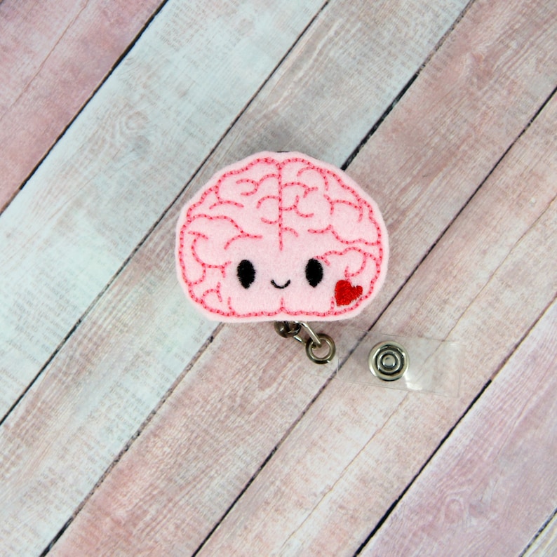 May include: A pink felt brain-shaped badge reel with a smiling face and a red heart. The badge reel has a retractable cord and a clear plastic holder for an ID or card. The background is a light wood grain.