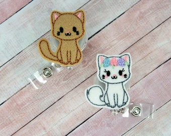 May include: Two embroidered cat badge reels. One is brown with a simple face and the other is white with a flower crown and a pink nose.