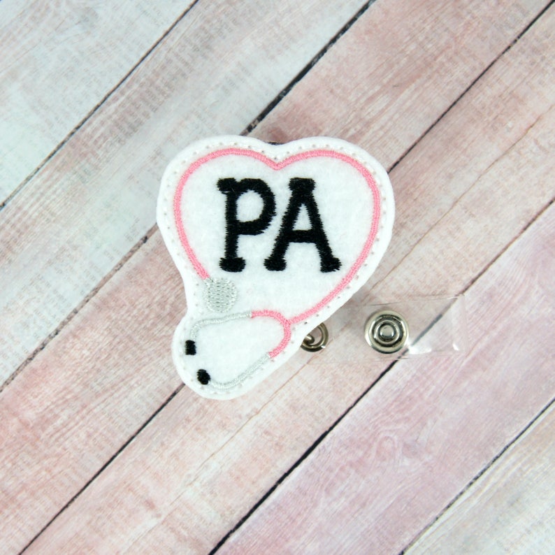 May include: White heart-shaped badge with a stethoscope and the letters "PA" in black embroidery. The badge is attached to a clear plastic retractable badge holder.