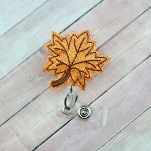 May include: A gold glitter maple leaf with a brown outline is attached to a clear plastic retractable badge holder.