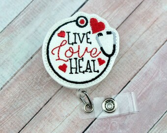 Könnte beinhalten: Runder weißer Badgehalter mit schwarzem Rand und roten gestickten Herzen. Der Badgehalter hat ein Stethoskop und den gestickten Text "Live Love Heal". Der Badgehalter ist an einem durchsichtigen, einziehbaren Badge-Band befestigt.