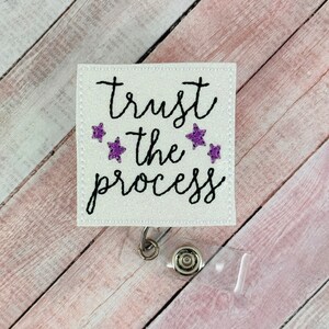 Może przedstawiać: Biały błyszczący uchwyt na identyfikator z tekstem "trust the process" w czarnym, ozdobnym pismem i czterema fioletowymi gwiazdami. Uchwyt na identyfikator jest przymocowany do przezroczystego plastikowego zwijacza.