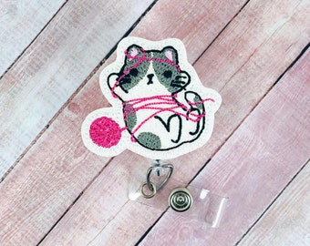 May include: A white embroidered badge with a gray and white cat tangled in pink yarn. The cat is holding a pink ball of yarn. The badge is attached to a clear plastic retractable badge holder with a silver clip.
