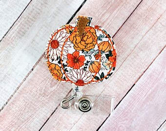 May include: A pumpkin-shaped badge reel with a floral design. The badge reel features a white background with orange and yellow flowers and green leaves. The pumpkin has a brown stem and is attached to a clear plastic reel with a metal clip.