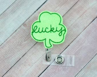 Puede incluir: Trébol de fieltro verde con la palabra "lucky" bordada en hilo verde. El trébol está unido a un portagafete de plástico transparente.