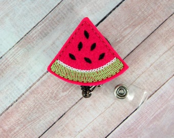 May include: A pink felt watermelon slice with black seeds and green embroidered edges attached to a clear plastic badge reel with a silver metal clip.