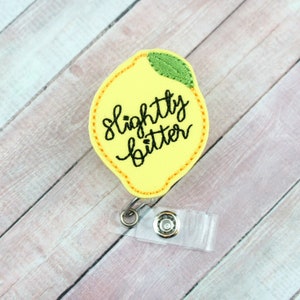 May include: Yellow lemon-shaped badge holder with the text "slightly bitter" embroidered in black. The badge holder has a clear plastic clip and a metal swivel.