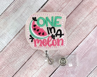 May include: A pink fabric badge holder with the text "ONE in a melon" and an embroidered watermelon design. The badge holder is attached to a clear plastic badge reel.