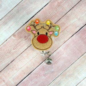 May include: A brown felt reindeer with a red nose and colorful lights around its antlers. The reindeer is attached to a clear plastic retractable badge holder.