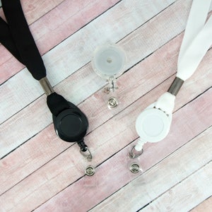 May include: Three retractable badge holders with lanyards. One is black, one is white, and the third is clear. Each has a metal clip and a clear plastic snap. The lanyards are made of a woven fabric.