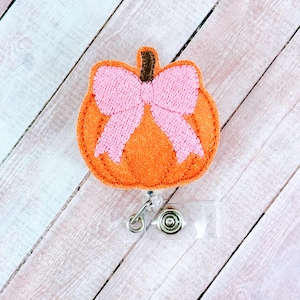 May include: Orange felt pumpkin with a pink felt bow, attached to a clear plastic retractable badge holder.