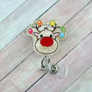 May include: Gold glitter reindeer with a red nose and colorful lights on its antlers. The reindeer is attached to a clear plastic retractable badge holder.