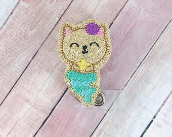 May include: A gold glitter cat with a purple flower on its head and a teal mermaid tail. The cat is holding a yellow star in its paws. The cat is attached to a clear plastic badge holder.