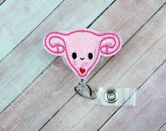 May include: Pink felt anatomical heart with a smiling face and a small red heart on the body. The heart is attached to a clear plastic retractable badge holder.