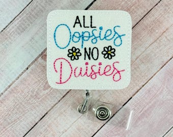 May include: White badge holder with the text "All Oopsies No Daisies" in pink, blue, and black embroidery. The badge holder has a clear plastic strap and a silver swivel clip.