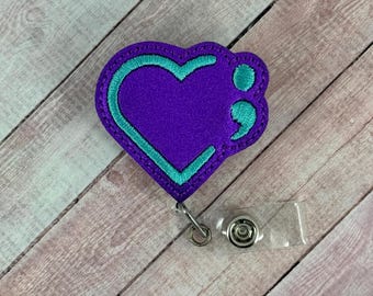 May include: Purple heart-shaped badge with a teal semicolon outline. The badge is attached to a clear plastic retractable badge holder.