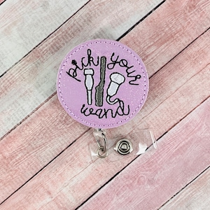 May include: A purple glitter badge holder with a white embroidered design that says "Pick your wand" with three wand images. The badge holder has a clear plastic retractable badge clip.
