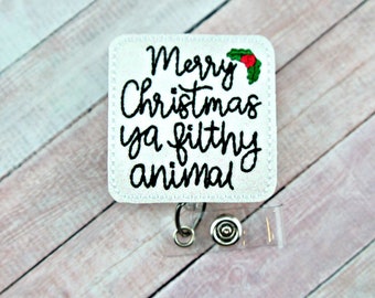 May include: White badge holder with a white background and black text that reads "Merry Christmas ya filthy animal". A small red and green holly sprig is embroidered above the text. The badge holder has a clear plastic strap and a silver clip.