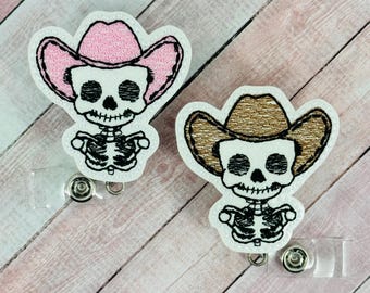 Puede incluir: Dos figuras de esqueleto bordadas con sombreros de vaquero. Un sombrero es rosa y el otro es marrón. Ambas figuras están unidas a soportes de credenciales retráctiles.