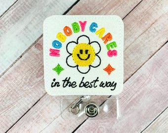May include: White embroidered badge reel with the text "NOBODY CARES" in rainbow colors, a smiling flower, and the phrase "in the best way." The reel has a clear plastic retractable cord and a metal clip.