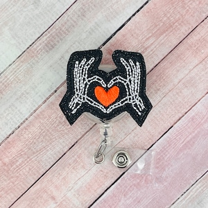 Puede incluir: Manos de esqueleto blancas y negras formando una forma de corazón con un corazón rojo en el centro. Las manos están unidas a un carrete de insignia de plástico transparente.