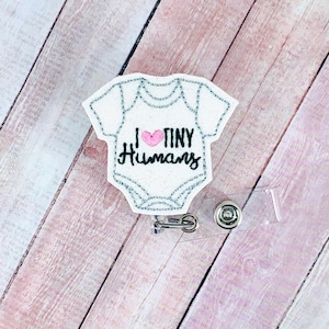May include: White fabric baby onesie with a pink heart and the text "I ♥ TINY HUMANS" embroidered on it. The onesie is attached to a clear plastic badge holder with a metal clip.