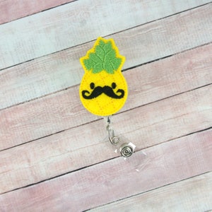 Op de afbeelding: Een gele vilt ananas met een zwarte vilt snor en groene vilt bladeren bevestigd aan een transparante, intrekbare badgehouder van plastic.