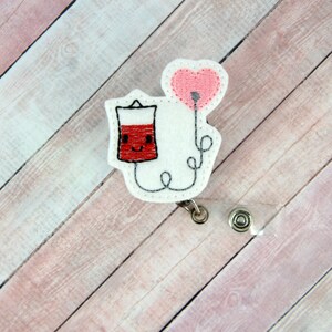 May include: White felt badge reel with a red and white cartoon blood bag with a smiling face and a pink heart balloon attached to the top. The blood bag is connected to the balloon by a white string.
