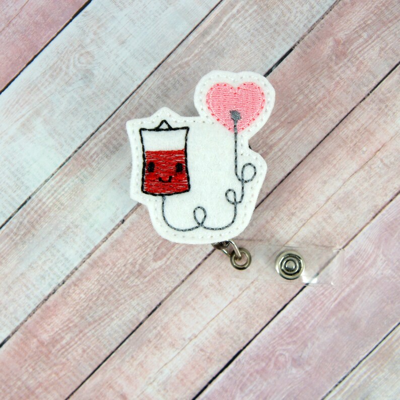 May include: White felt badge reel with a red and white cartoon blood bag with a smiling face and a pink heart balloon attached to the top. The blood bag is connected to the balloon by a white string.