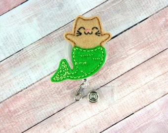 May include: A green and brown felt cat mermaid badge reel. The cat has a smiling face and is wearing a green mermaid tail. The badge reel is attached to a clear plastic retractable cord.