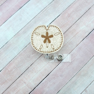 May include: A white glitter sand dollar with a brown embroidered starfish design. The sand dollar is attached to a clear plastic retractable badge holder with a silver clip and button.