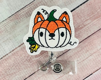 May include: A white embroidered patch with a fox face inside an orange pumpkin. The patch is attached to a clear plastic retractable badge holder with a silver clip and a silver swivel.