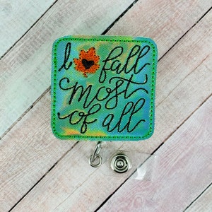 以下が含まれることがあります： 「I fall most of all」というフレーズが黒い筆記体で書かれた正方形のバッジリール。背景は青と緑の色合いの水彩デザインで、緑色のステッチの縁取りが施されています。黒いハートの付いたオレンジ色の葉が左上にあります。