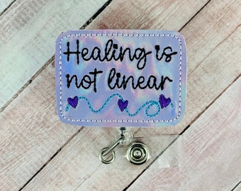 May include: A rectangular badge reel with the text "Healing is not linear" in black script. The badge has a holographic, iridescent background with a stitched border. A teal wavy line with purple hearts decorates the bottom.