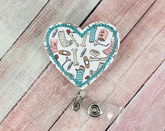 May include: A white heart-shaped badge holder with a teal blue stitched border. The holder features a pattern of medical supplies, including bandages, casts, and crutches.