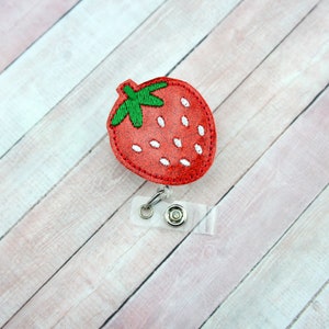 May include: Red glitter strawberry with green leaves and white seeds, attached to a clear plastic retractable badge holder.