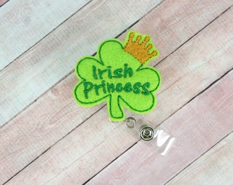 Puede incluir: Trébol de fieltro verde con una corona dorada y el texto "Irish Princess" en él. El trébol está unido a un portagafete retráctil de plástico transparente.