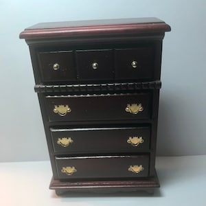 May include: A dark brown wooden chest of drawers featuring three small top drawers with round silver knobs and three larger lower drawers with gold-toned metal swing handles. The piece has a flat top surface and a decorative horizontal trim.
