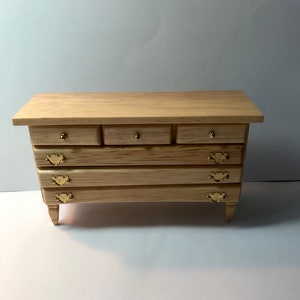 May include: A miniature wooden dresser featuring a light tan wood grain finish. The piece includes three small top drawers with round gold knobs and three larger lower drawers, each equipped with two decorative gold-toned metal swing handles.
