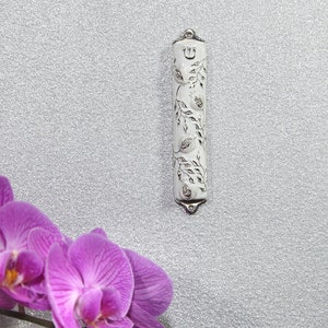 May include: A white metal Torah pointer with a silver design of pomegranates and leaves. The pointer has a silver Hebrew letter "Shin" at the top.