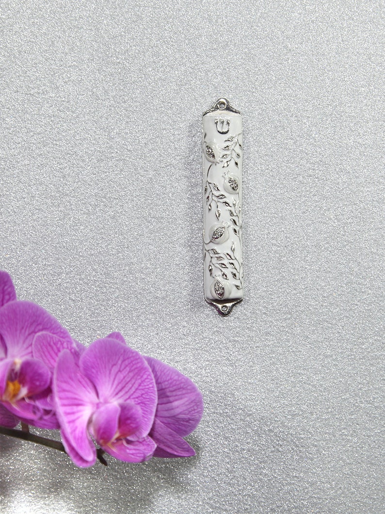 May include: A white metal Torah pointer with a silver design of pomegranates and leaves. The pointer has a silver Hebrew letter "Shin" at the top.