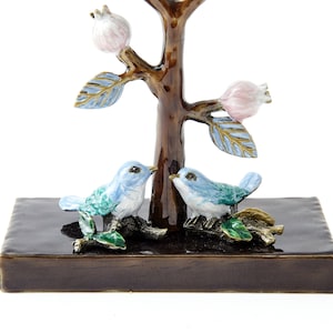May include: A decorative tree figurine with two blue birds perched on branches. The tree has a brown trunk and pink blossoms. The birds are facing each other and have green accents. The tree is sitting on a dark brown base.