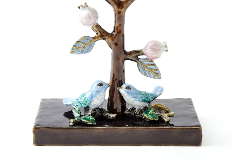 May include: A decorative tree figurine with two blue birds perched on branches. The tree has a brown trunk and pink blossoms. The birds are facing each other and have green accents. The tree is sitting on a dark brown base.