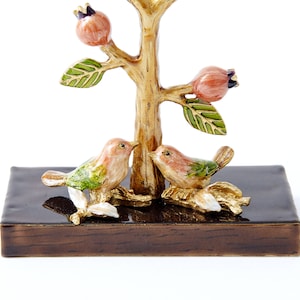 May include: A gold-tone metal jewelry tree with two birds perched on branches. The tree has a brown wooden base and pink flowers.
