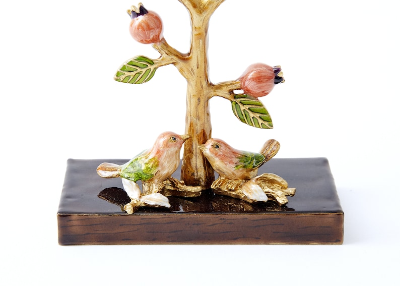 May include: A gold-tone metal jewelry tree with two birds perched on branches. The tree has a brown wooden base and pink flowers.