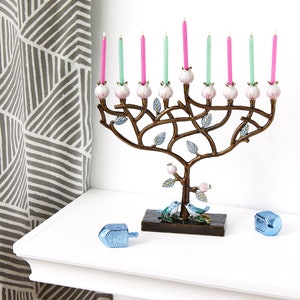 May include: A menorah shaped like a tree with pink and green candles. The menorah is decorated with small white flowers and blue birds. Two blue dreidels are on a white surface in front of the menorah.