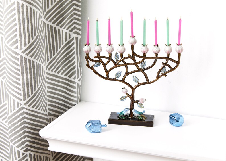 May include: A menorah shaped like a tree with pink and green candles. The menorah is decorated with small white flowers and blue birds. Two blue dreidels are on a white surface in front of the menorah.