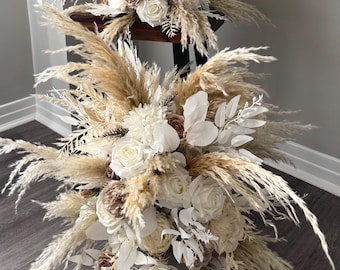 Op de afbeelding: Twee lagen bloemstuk met pampasgras, witte rozen en accenten van bruine en witte bloemen. Het arrangement wordt gepresenteerd op een donkere houten tafel, wat een neutrale en elegante uitstraling creëert.