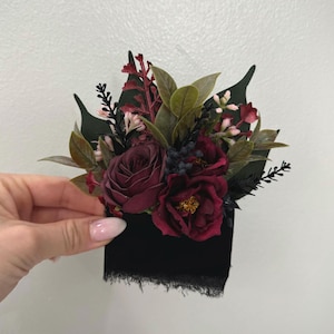 May include: A dark burgundy floral arrangement featuring roses, berries, and greenery. The flowers are set against a black base with a frayed edge. The arrangement is held against a white wall.